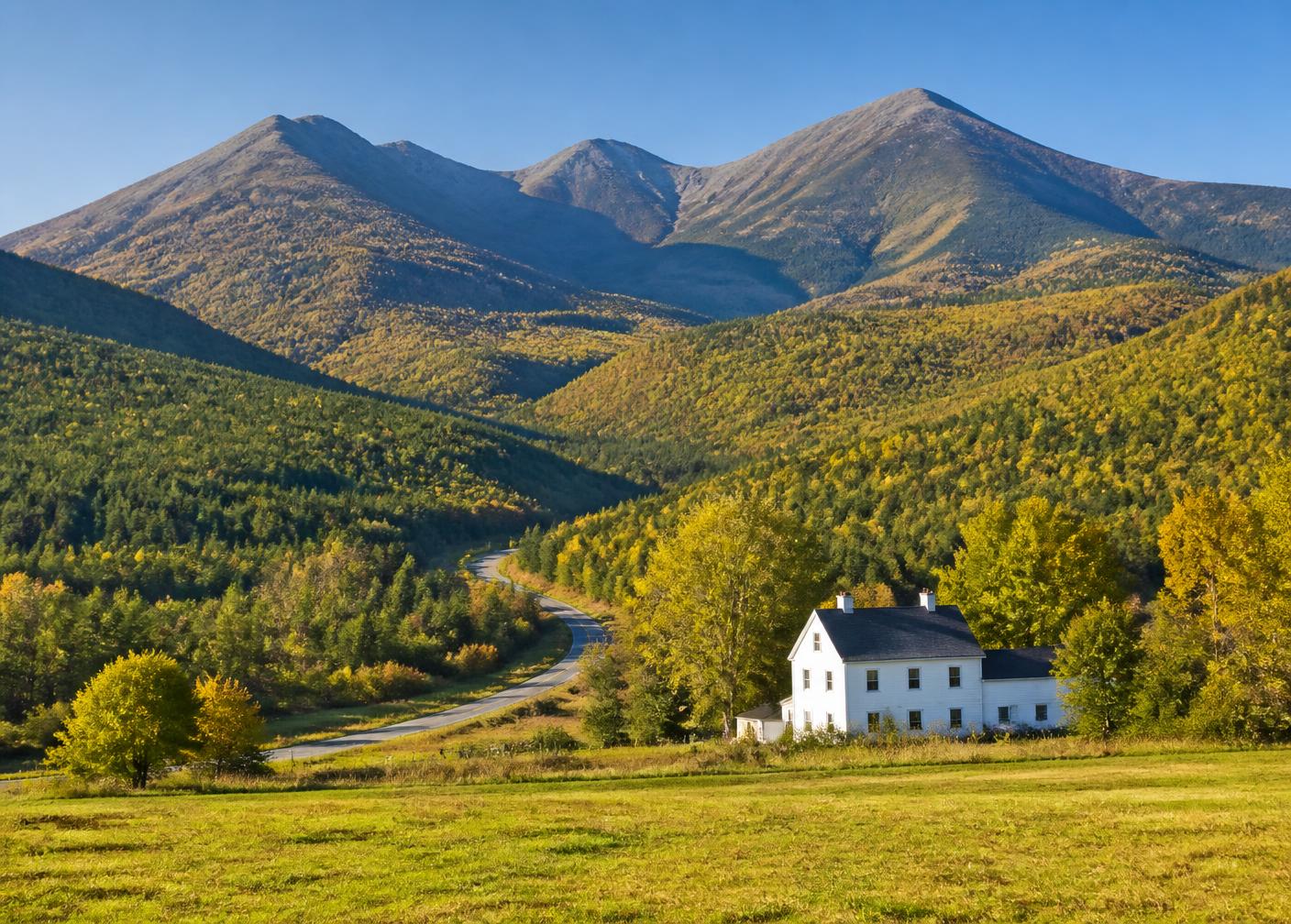 White farmhouse in a green meadow below the forested peaks of the White Mountains in early autumn
